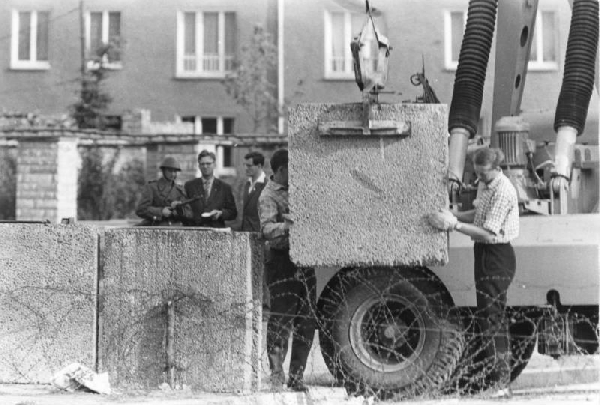 Falépítés Berlinben; 1961. augusztus. A kép előterében megfigyelhetők a határzárás első óráiban kihúzott szögesdróttekercsek, a Fal 1. generációs változatára jellemző, kb. 1 m˛-es (eredetiben barnás színű) falazóelemek. A háttérben balra az NDK Néphadseregének egyik katonája (sisakban és PPS-41 géppisztollyal). A képen látható objektumok mind NDK területen vannak, a felvétel nyugati oldalról készült.